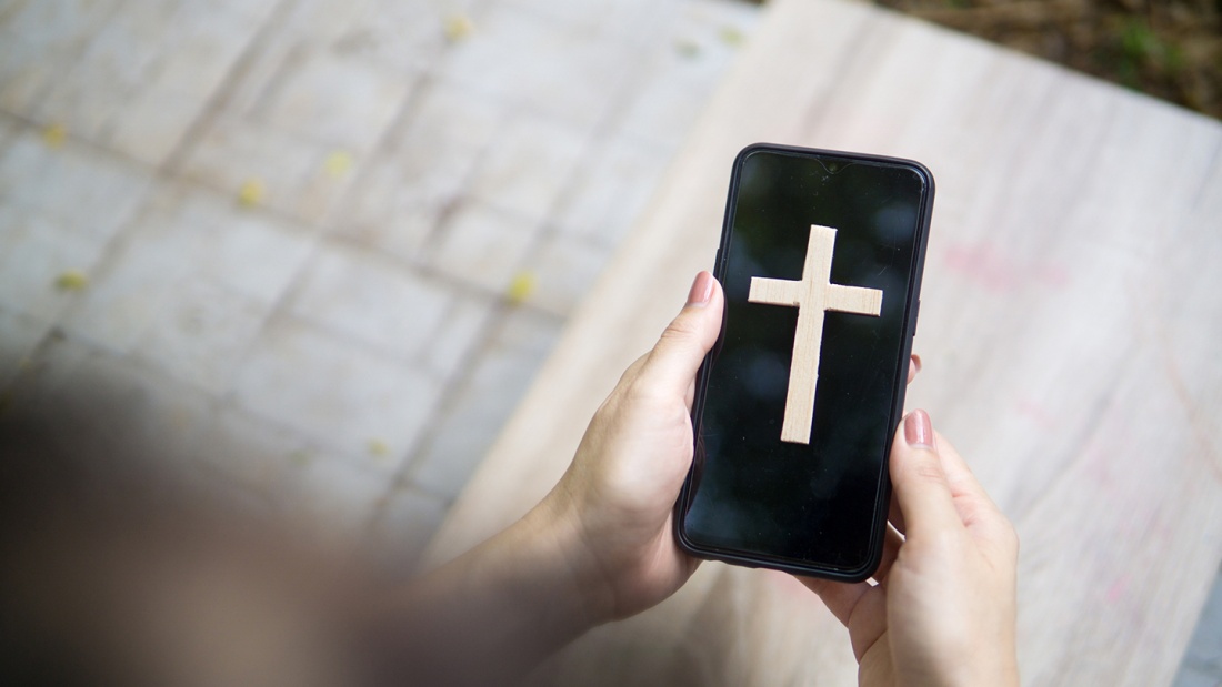 Frau hält Smartphone mit einem Kreuz drauf in der Hand.